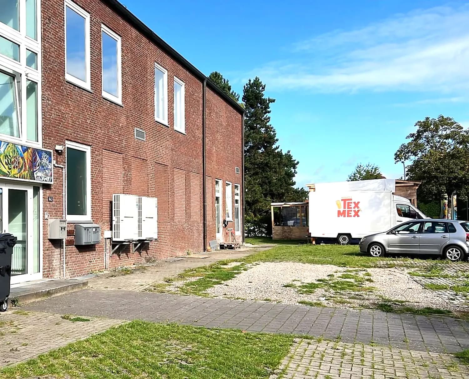 Rotes Backsteingebäude mit der Hausnummer 143, rechts daneben der weisse Wagen auf dem Platz