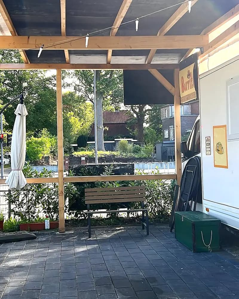 Blick unter die Holzpergola auf die Bank und den weissen Wagen daneben