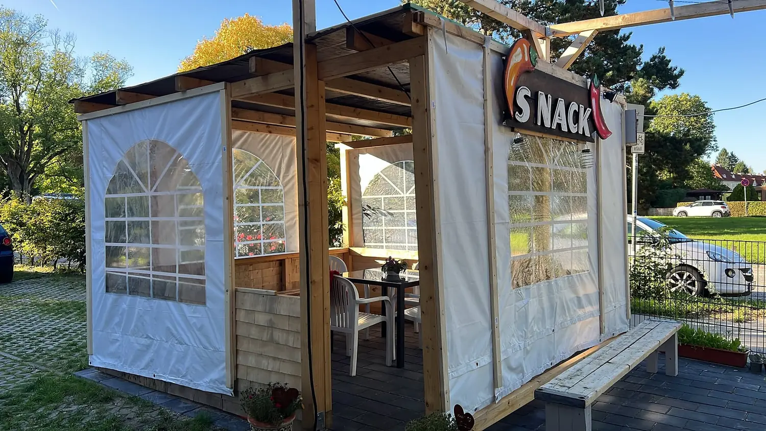 Überdachter Sitzplatz aus Holz mit weissen Planenwänden und Bogenfenstern, drinnen Tisch und Stühle, davor eine lange Holzbank, oben das Schild SNACK mit zwei Chilischoten