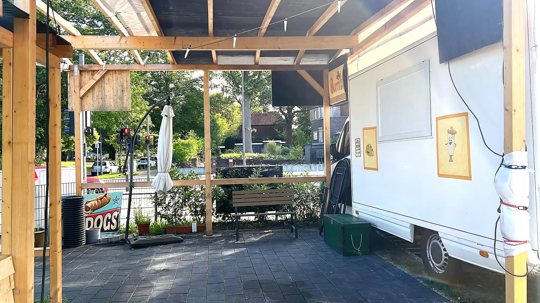 Überdachte Holzterrasse neben dem weissen Wagen: Pergola mit Lichterkette, eine Holzbank, ein zusammengebundener Sonnenschirm, dahinter die Strasse