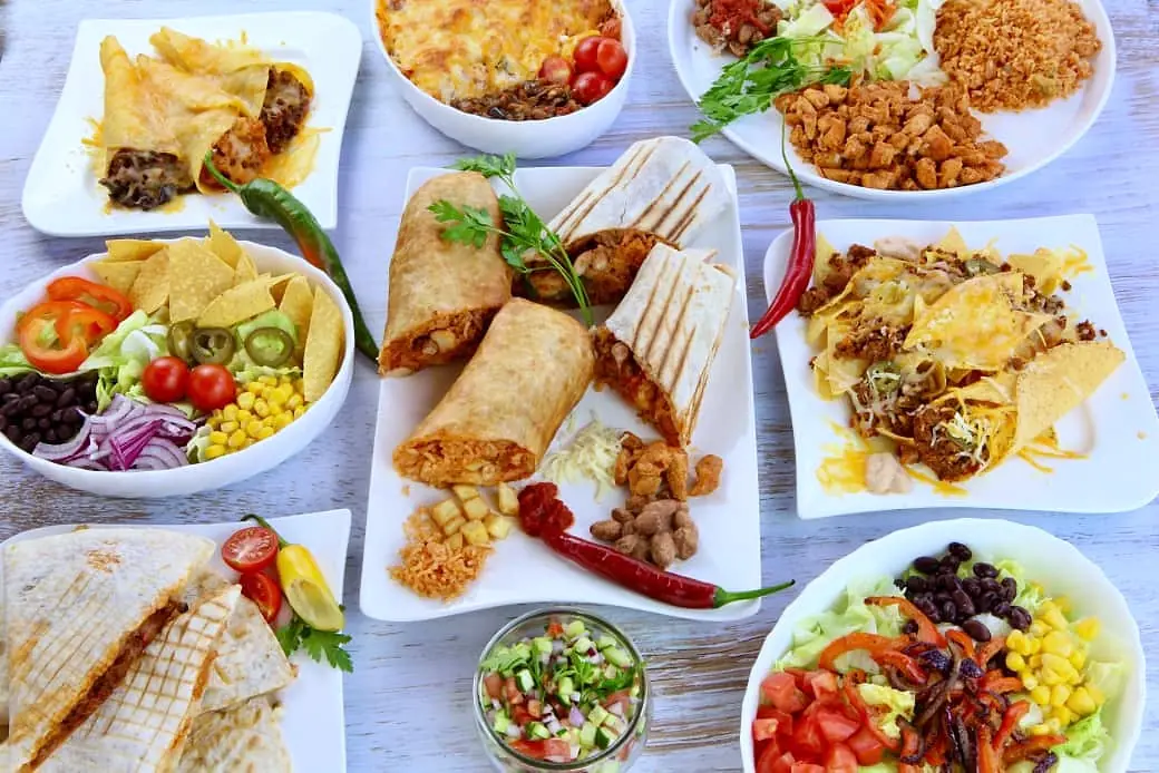 Gedeckte Tafel von oben auf hellem Holztisch: Burritos, Quesadillas, Taquitos, überbackene Nachos, Salatschalen mit schwarzen Bohnen und Mais, Tex-Mex-Reis und Salsa