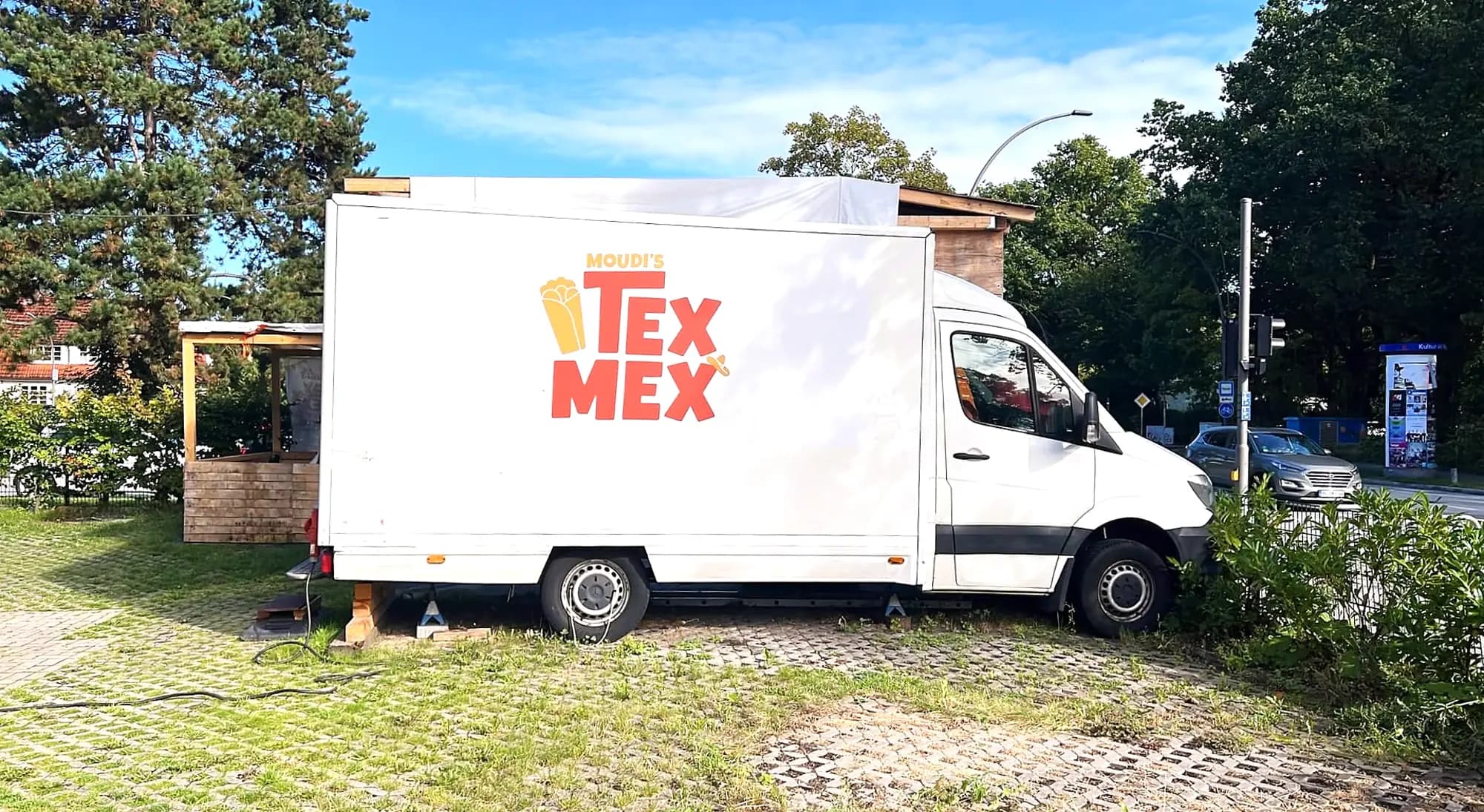 Der weisse Foodtruck von MOUDI'S TEX MEX mit dem Logo auf der Seitenwand, dahinter die überdachte Holzterrasse